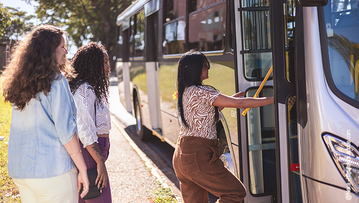 Passengers standing next to a waiting bus that has it door open. First person in line is stepping on.