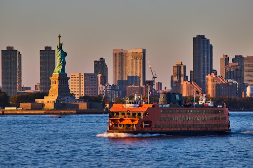 Large Staten Island Ferry passing by Statue of Liberty in golden hour of New York City with skyline behind