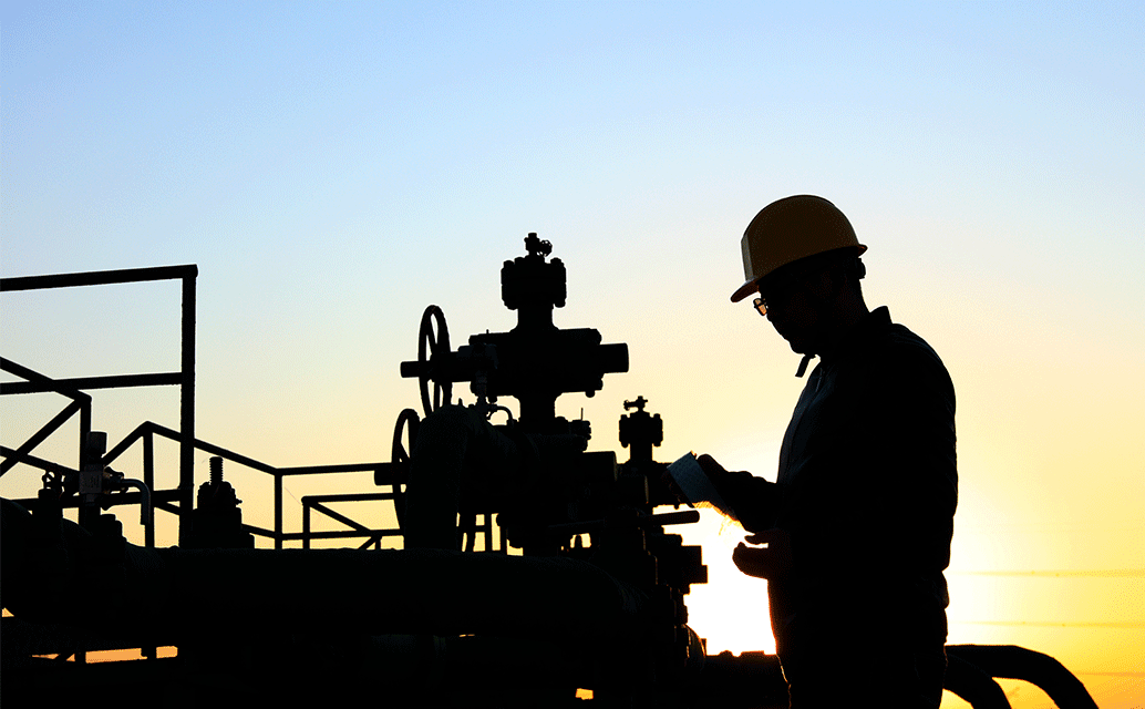 An image of an oil worker and oil pipes in silhouette in front of rising or setting sun.